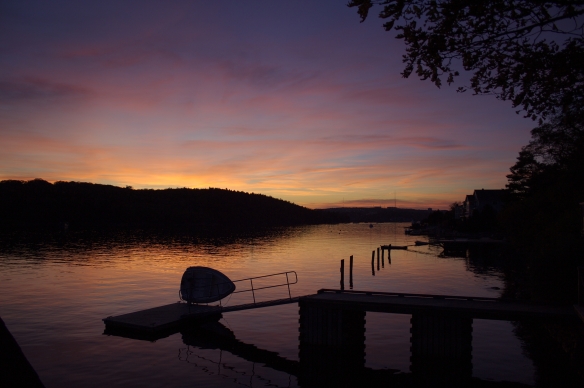 A photograph of a sunset over the northwest arm of Halifax harbour. In the foreground can be seen the silhouette of a private dock to which is attached a small boat, held completely out of the water, and turned on its side. The sky is dark blue or purple at the edges, with warm yellow-orange tones getting more and more intense toward the horizon. 